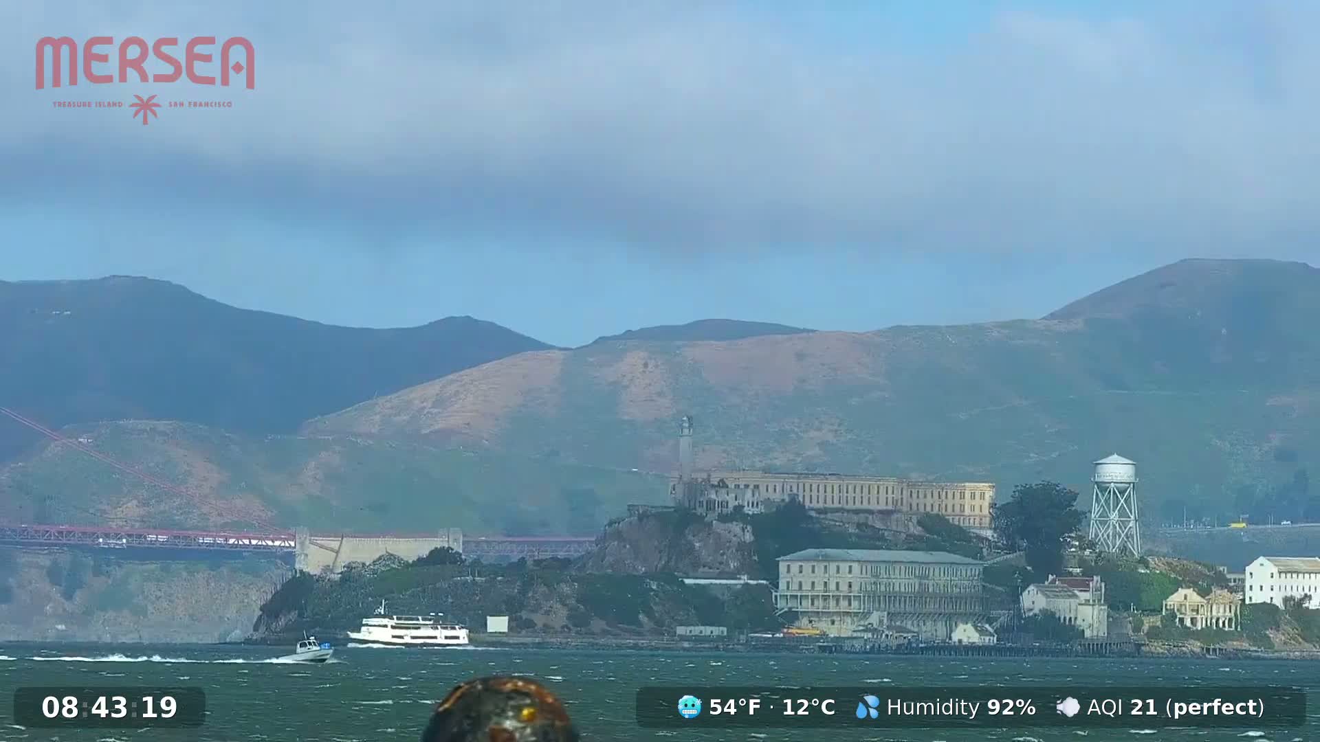 A hazy day shows Alcatraz Island with a ferry and other boats in the water, mountains in the background, and a portion of the Golden Gate Bridge visible on the left.
