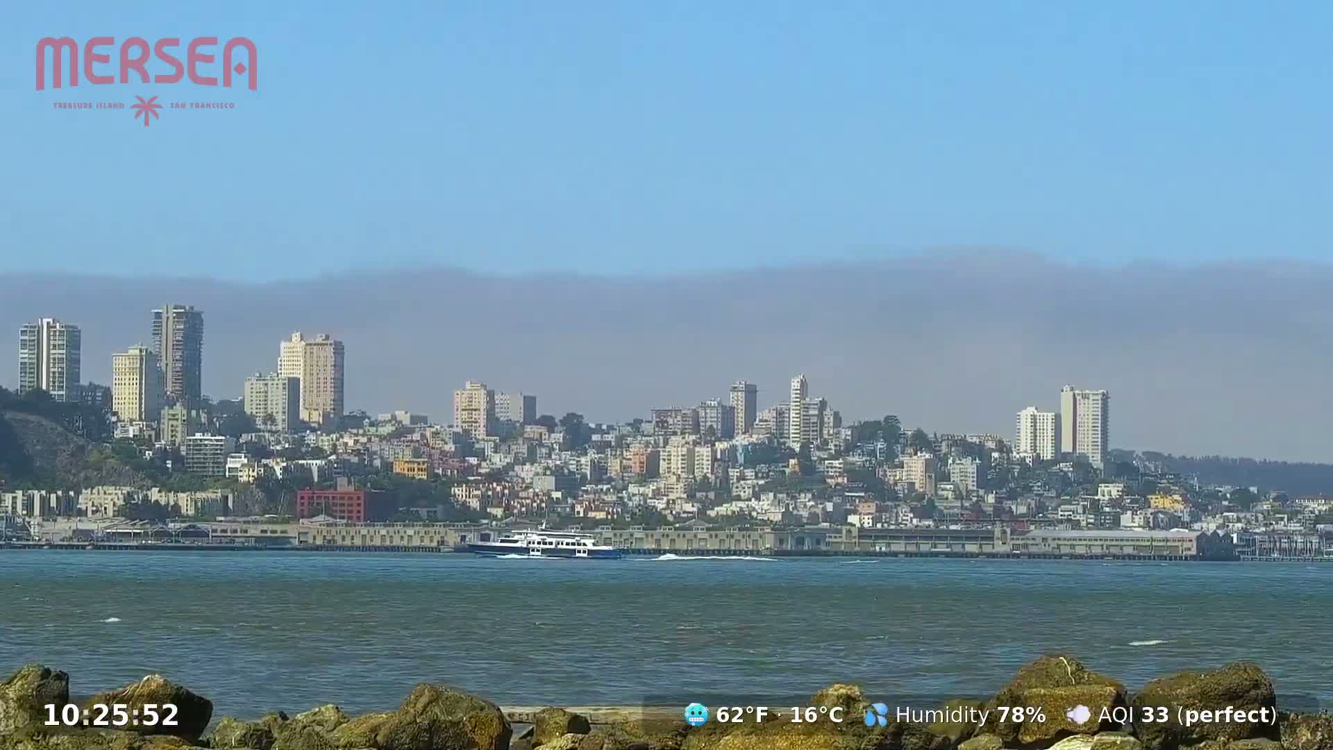 A hazy cityscape sits across a body of water, viewed from a low vantage point near some rocks.