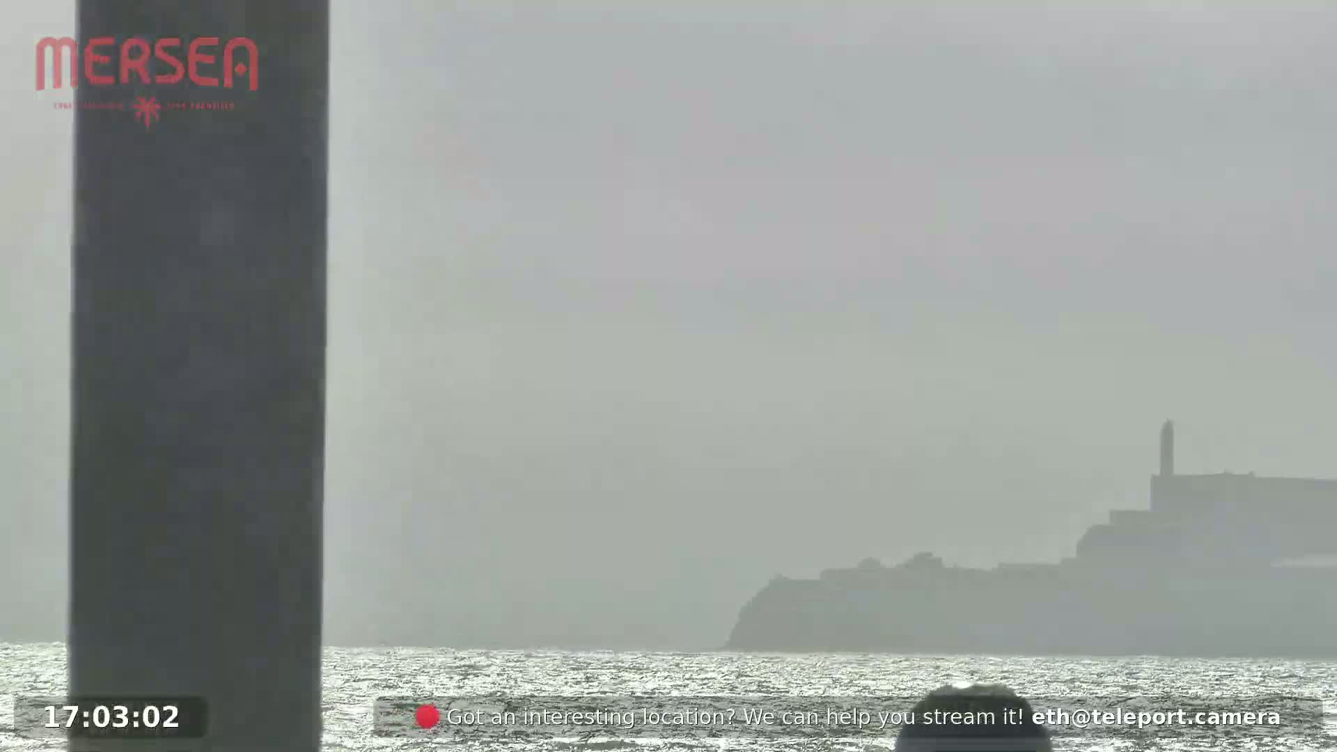 A hazy, gray day shows a distant island with a tall structure on the horizon, viewed from across a body of water.