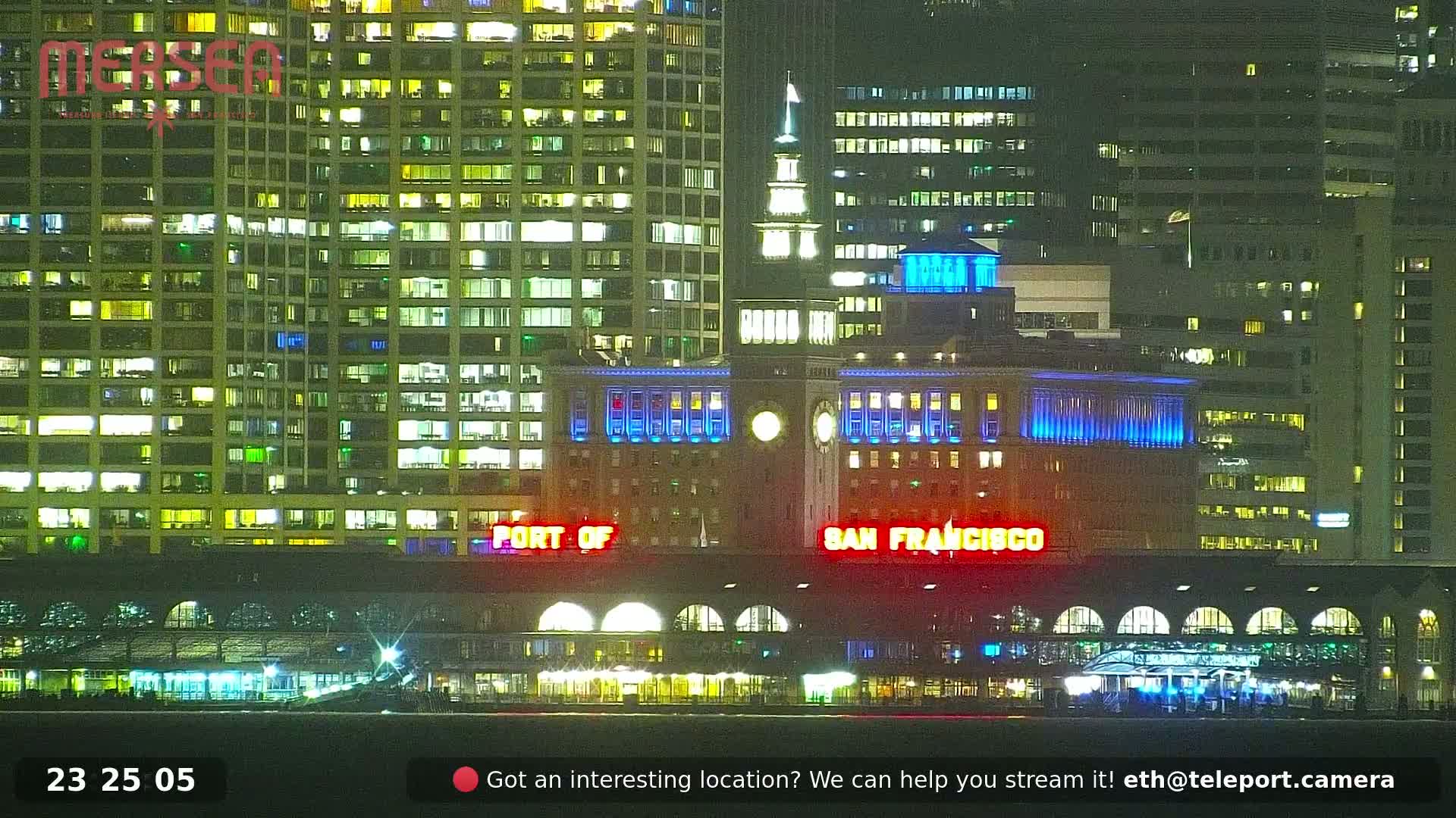 Nighttime view of brightly lit San Francisco buildings along a waterfront, with calm water in the foreground.