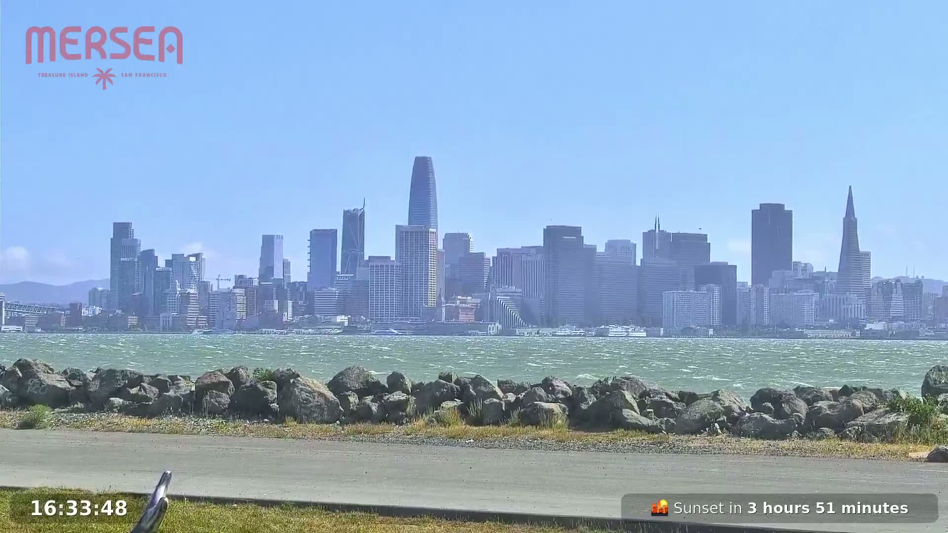 A city skyline is visible across a choppy body of water from a rocky shoreline on a sunny day.