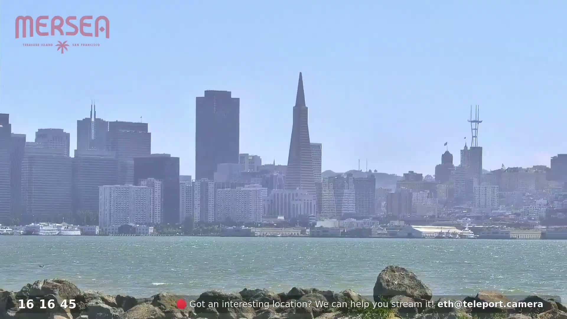 A hazy, sunny day shows a San Francisco cityscape across a body of water from a rocky shoreline.