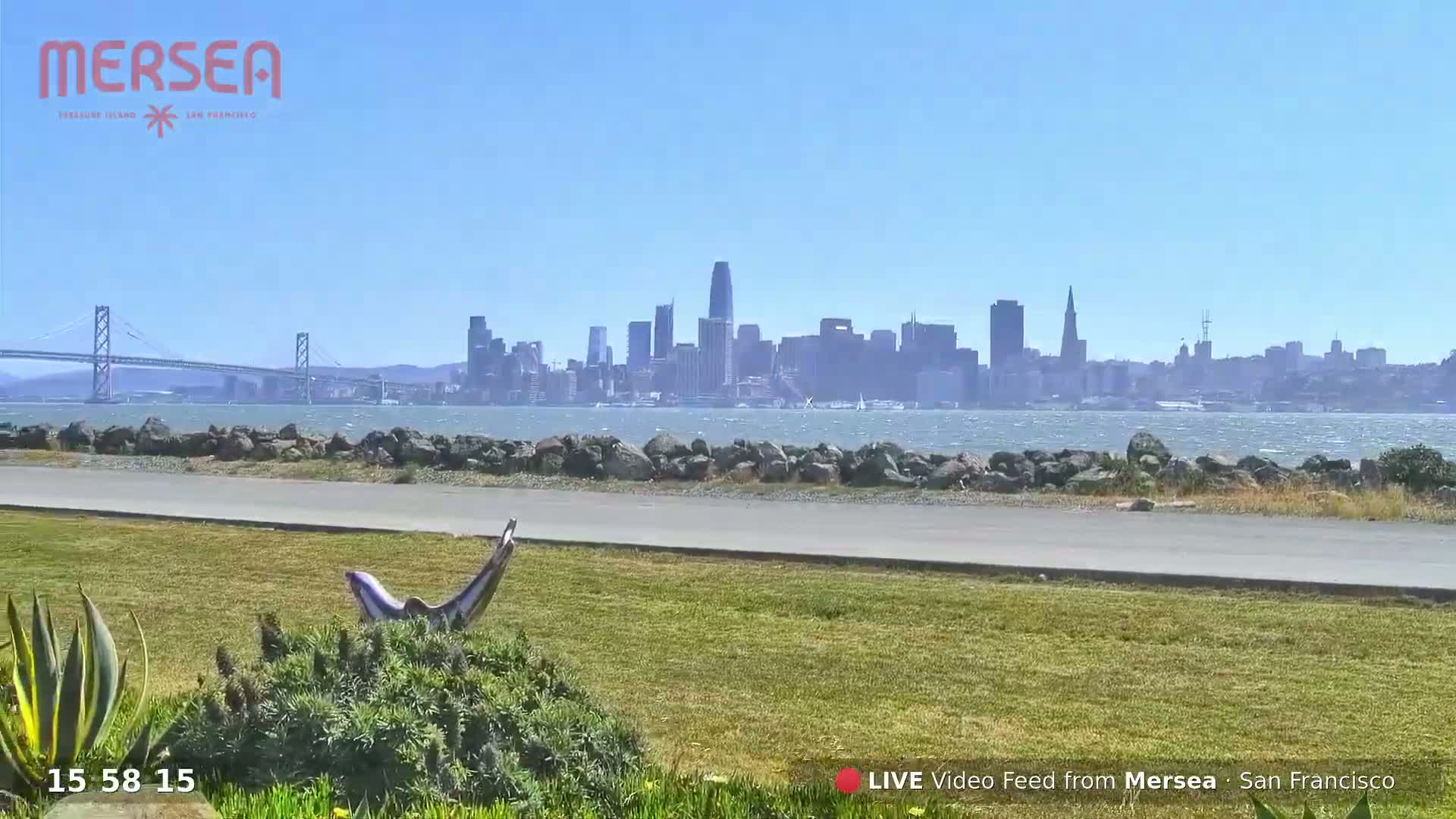 A grassy area with a small sculpture overlooks a body of water, a bridge, and the San Francisco skyline under a clear, sunny sky.