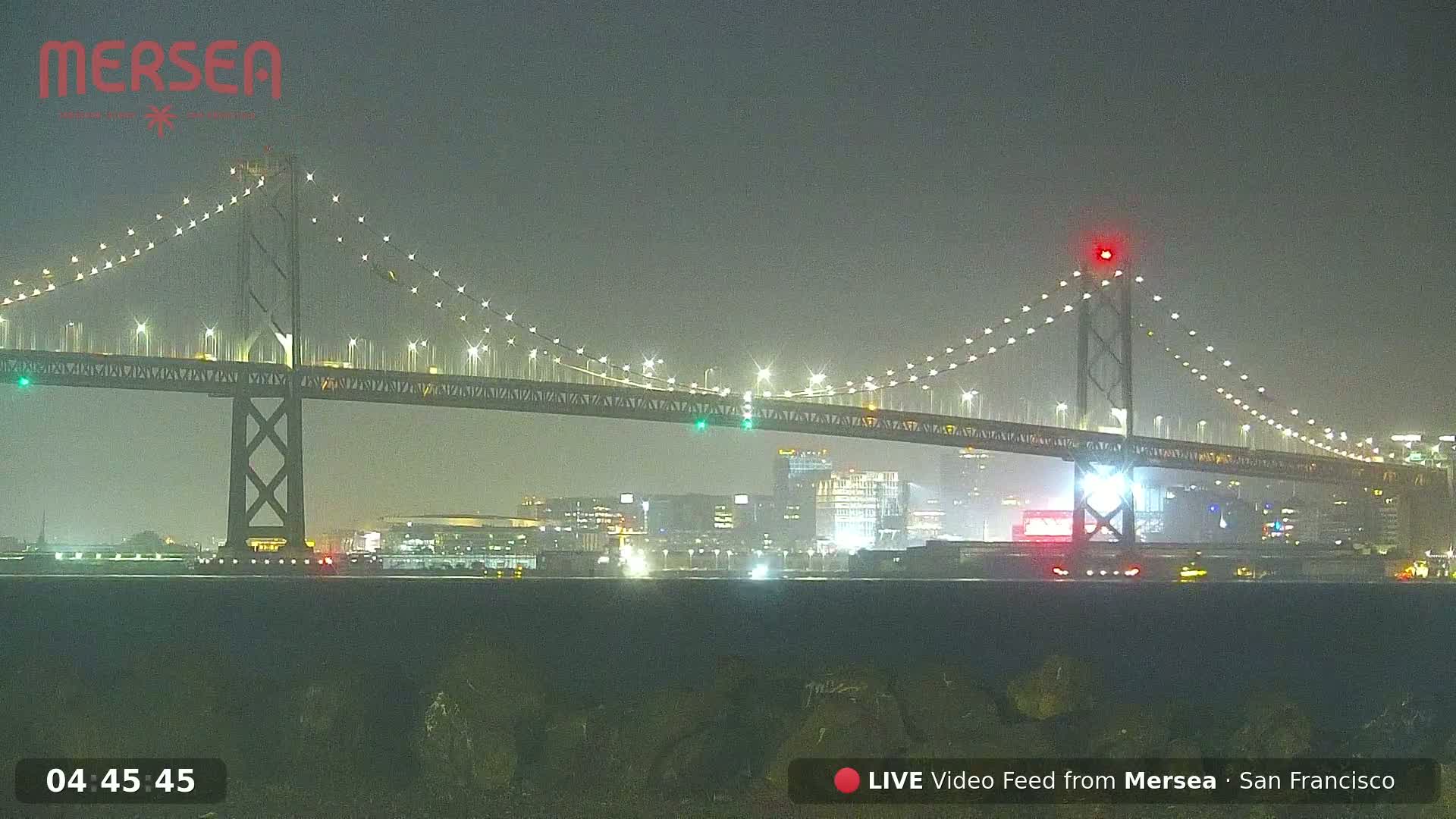 A lit suspension bridge is seen at night in hazy conditions over a cityscape.