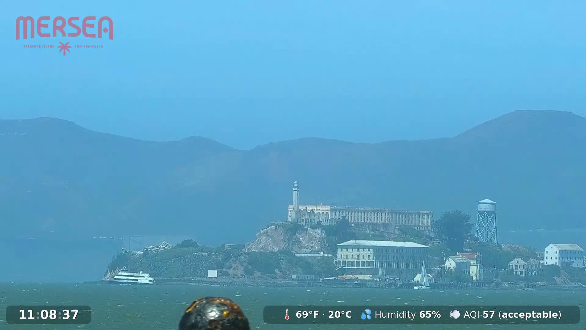 Alcatraz Island, with its buildings and lighthouse, is visible across calm water under a hazy blue sky.