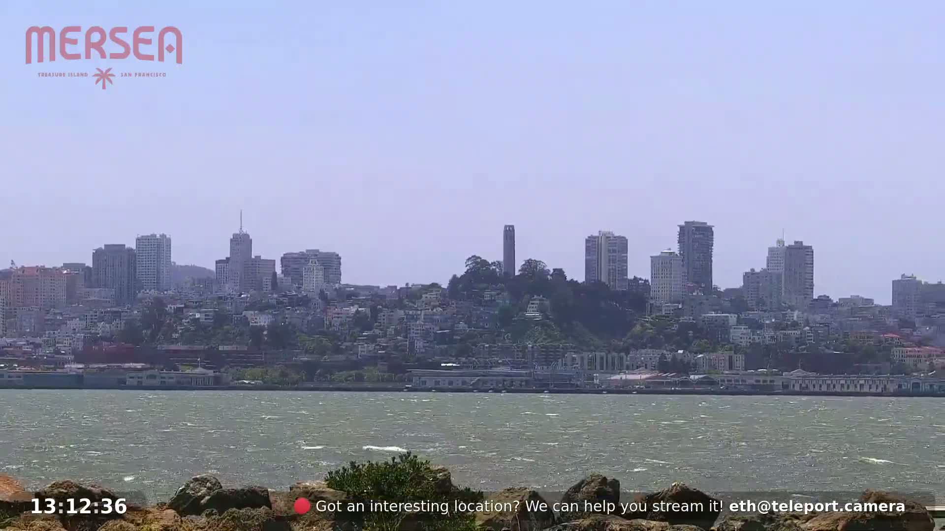 A cityscape with a tall tower on a hill is seen across a body of water under a hazy sky.