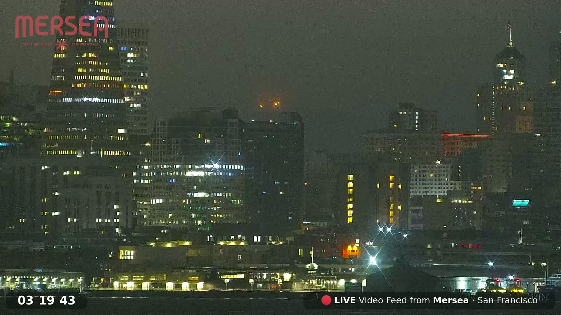 A nighttime cityscape of numerous illuminated buildings along a waterfront under hazy conditions.