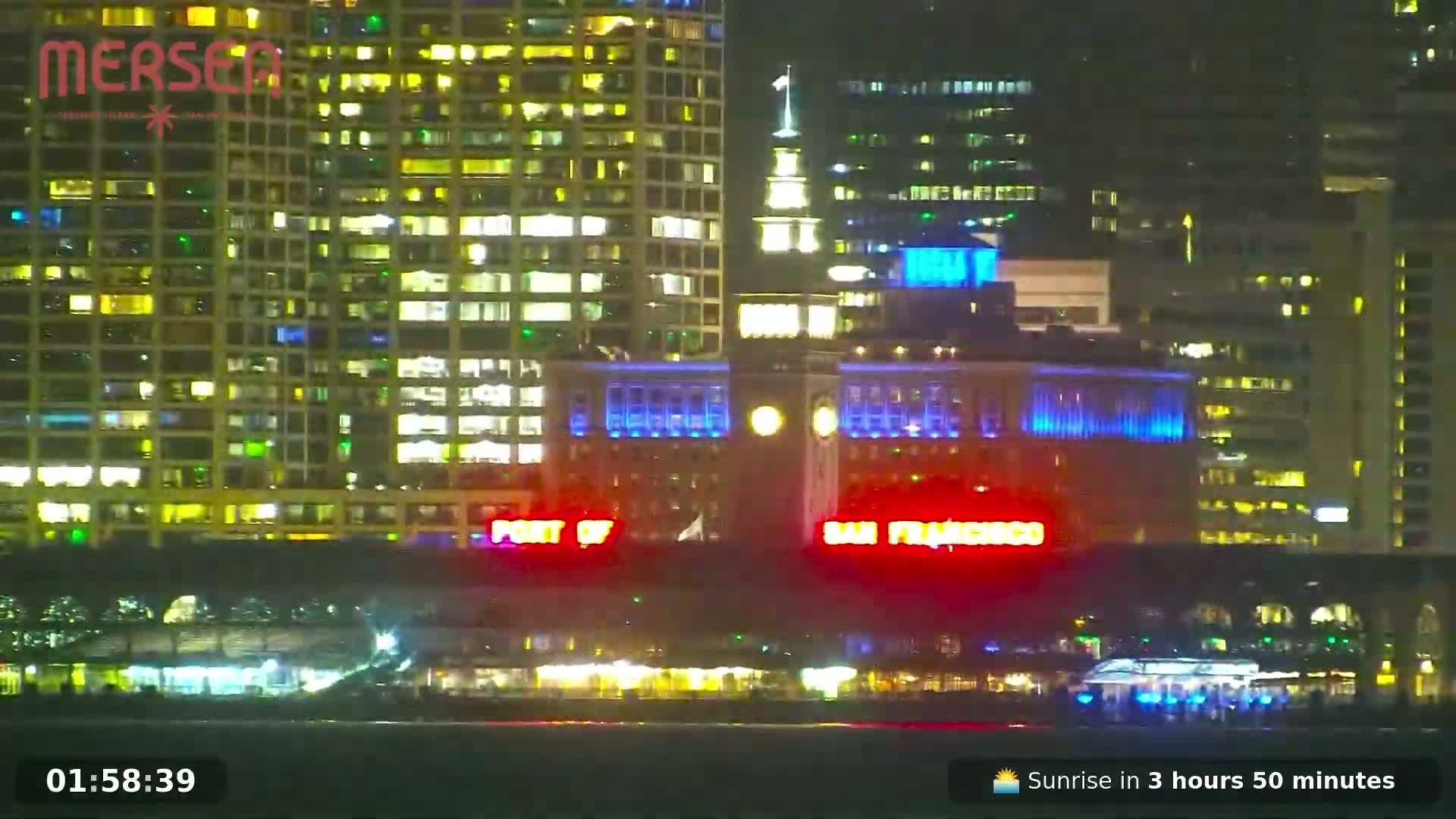 A nighttime view of brightly lit city buildings along a waterfront, with boats visible in the water.
