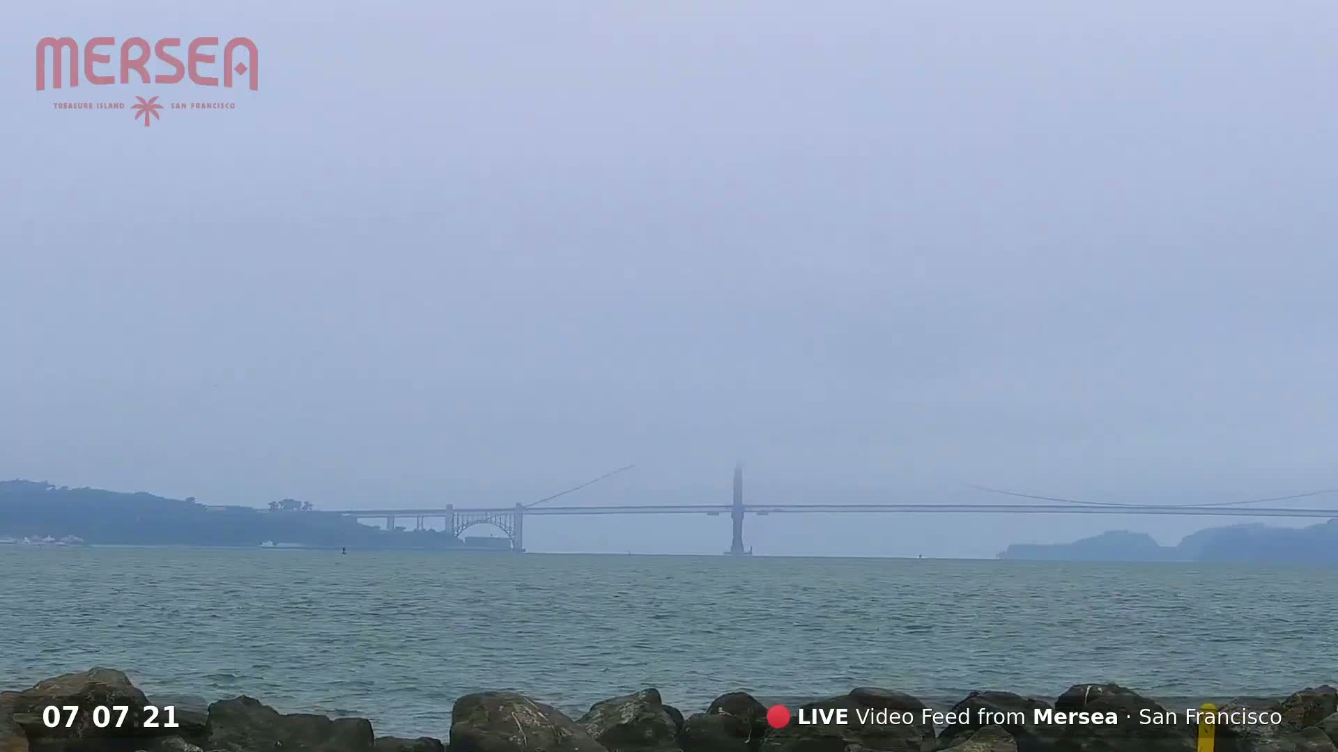 A hazy, overcast day shows a long bridge spanning a body of water, with dark rocks in the foreground.