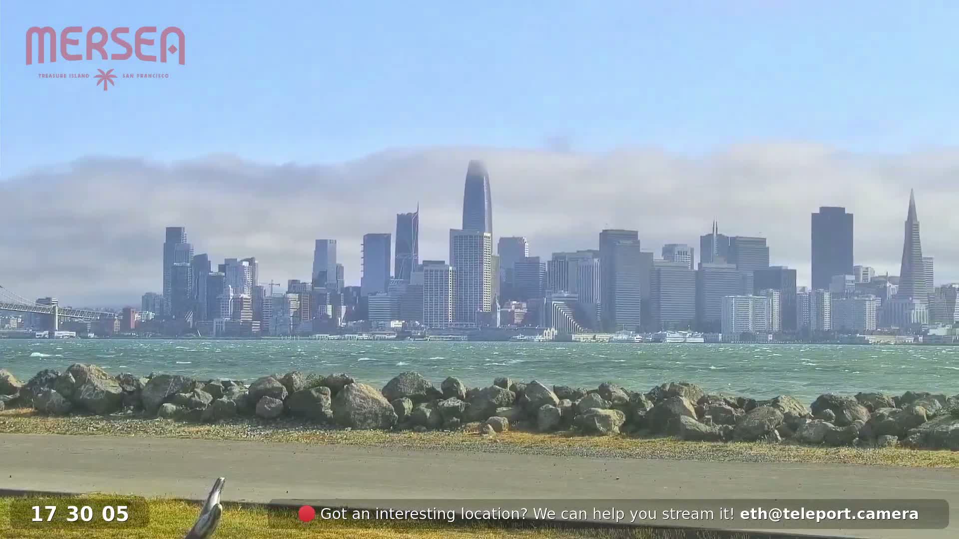 A hazy, windy day shows a city skyline across a body of water, with a rocky shoreline in the foreground.
