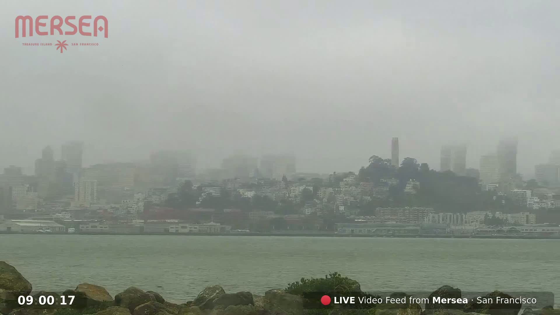 A foggy city skyline is seen from across a body of water, with dark rocks in the foreground.