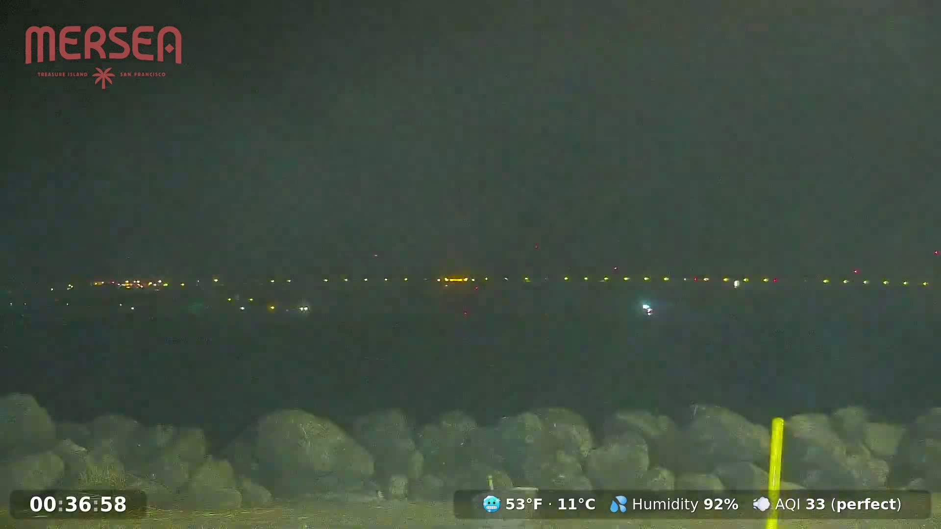 A nighttime view of a distant runway illuminated with lights, observed from a rocky, dark foreground under a hazy sky.