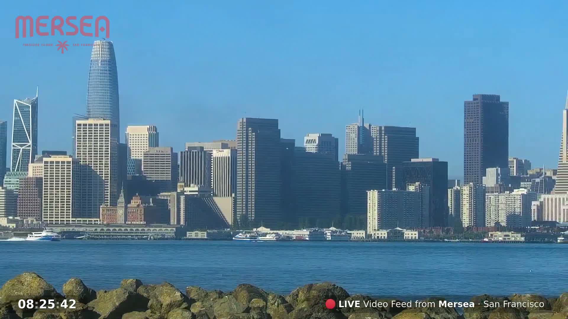 A clear, sunny day shows a cityscape with numerous skyscrapers and high-rise buildings along a waterfront, viewed from across a body of calm water.