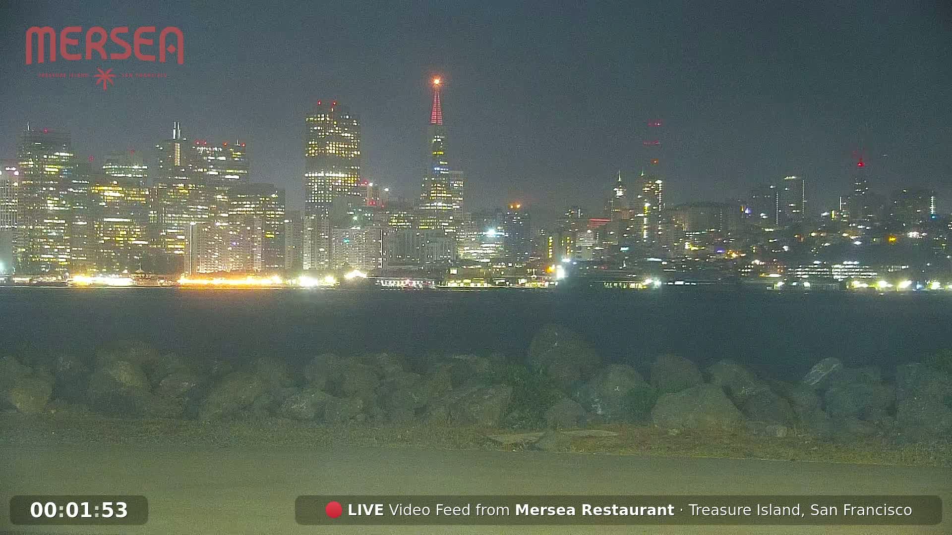 A nighttime view of a city skyline across a body of water, with a rocky shoreline in the foreground under a dark sky.