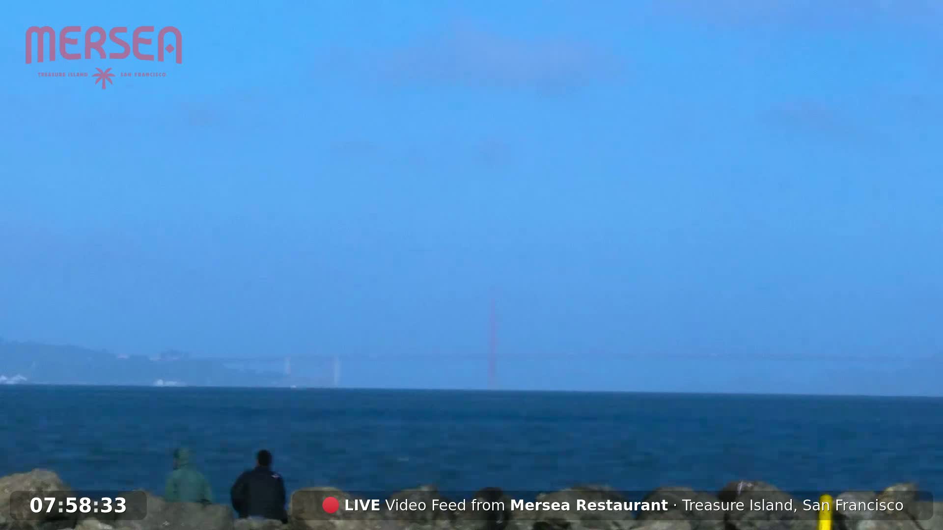 Two people stand on a rocky shore overlooking a calm ocean with a hazy view of a distant bridge under a clear, light-blue sky.