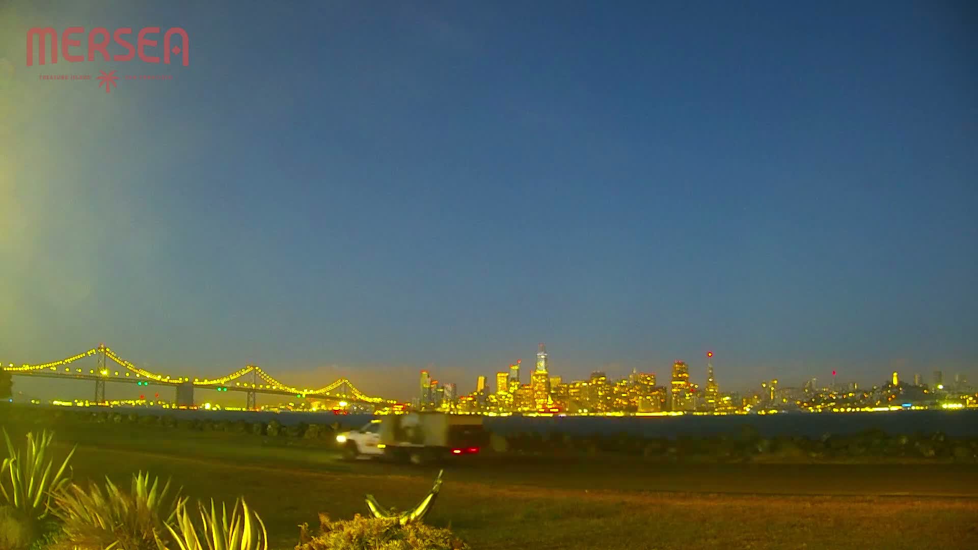 A brightly lit city skyline is visible across a body of water at dusk, with a bridge and a vehicle in the foreground.