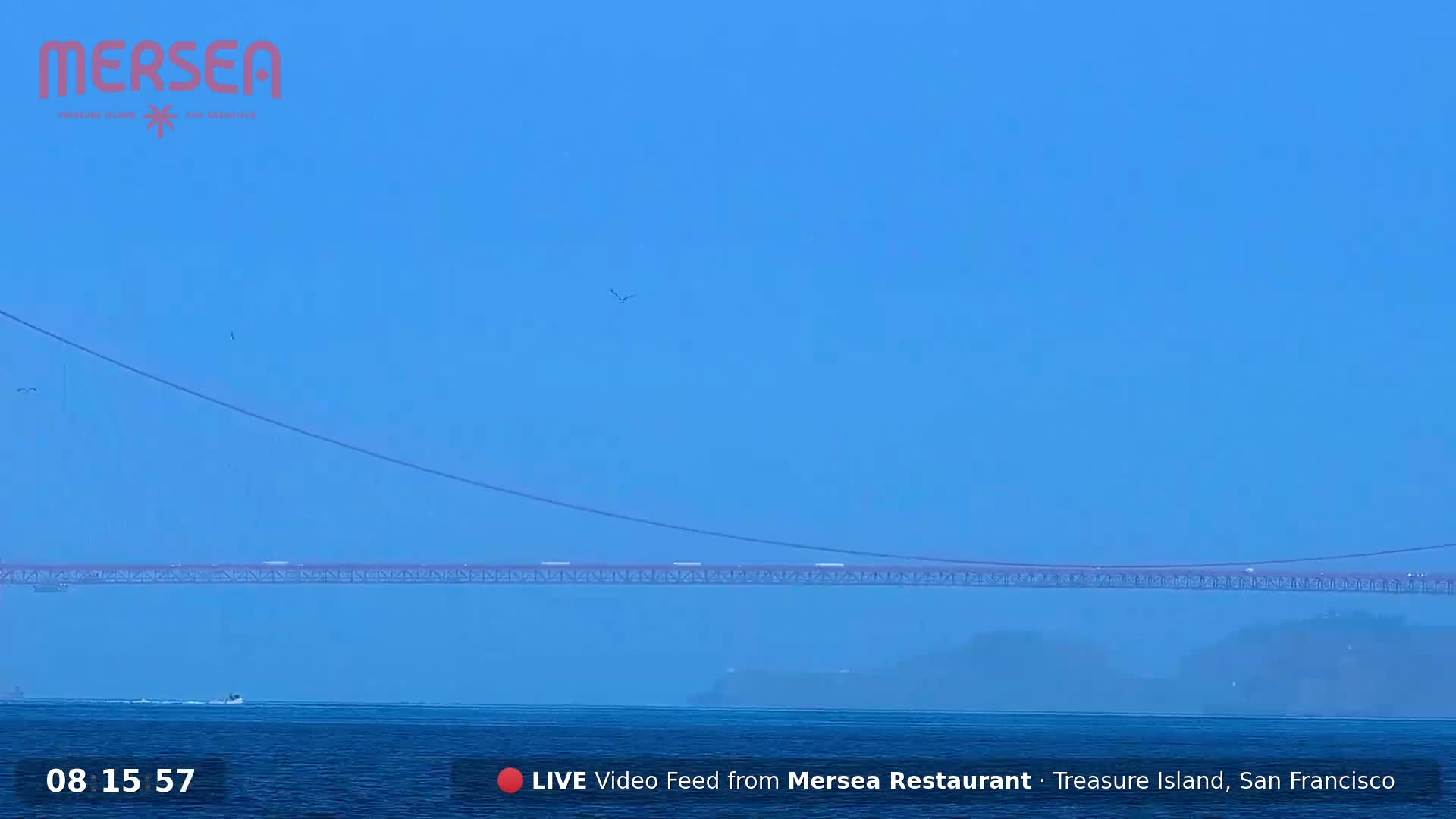 A long red bridge spans a hazy blue body of water under a clear blue sky.