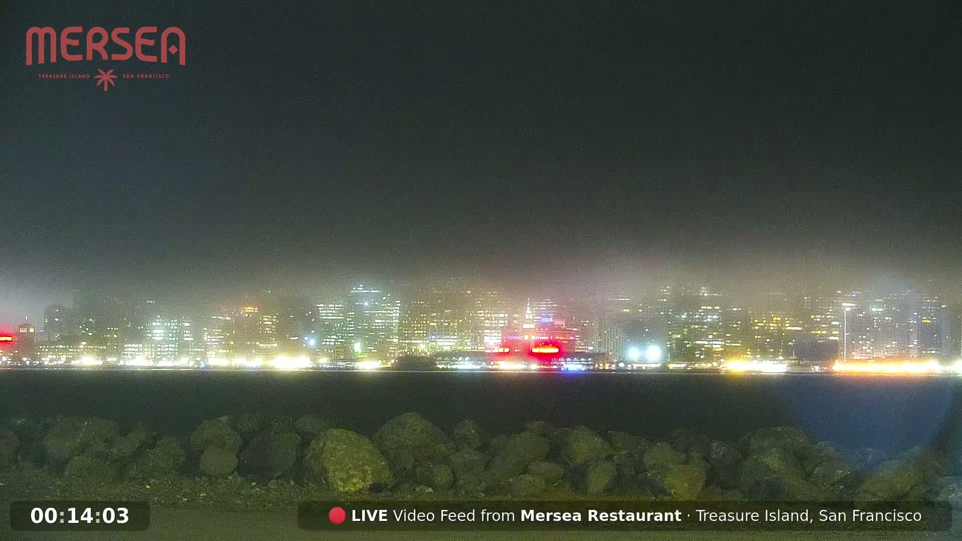 A foggy nighttime view of a city skyline illuminated by lights across a body of water, seen from a rocky shoreline.