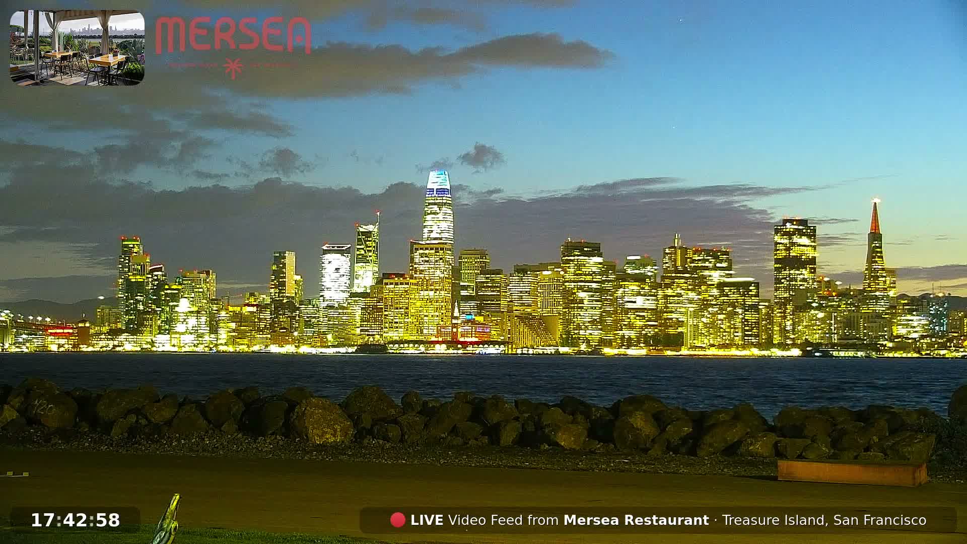 The San Francisco skyline, brightly illuminated at twilight across the bay with landmarks such as the Salesforce Tower and Transamerica Pyramid, is visible under a partly cloudy sky.