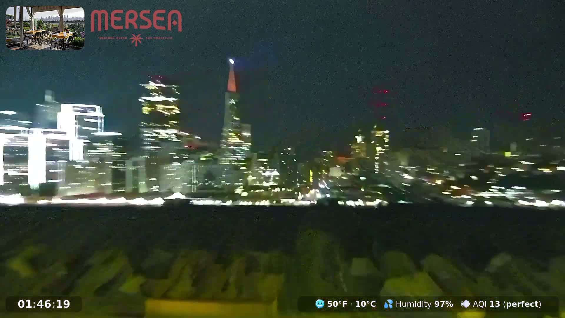 A blurry nighttime cityscape is illuminated by numerous building lights, including a prominent pyramid-shaped skyscraper, under outdoor conditions of 50°F (10°C) and 97% humidity.