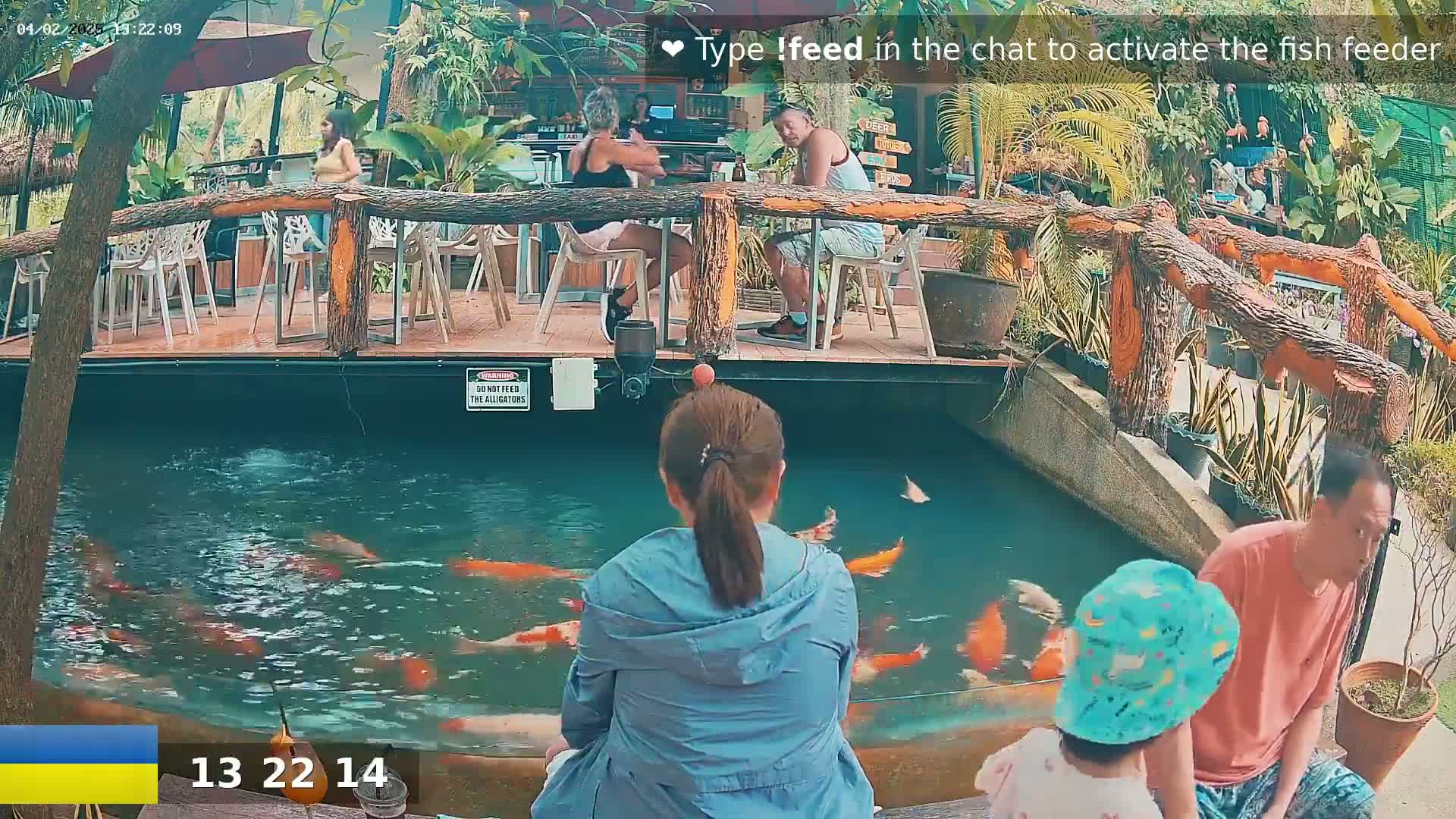A woman sits facing a pond filled with orange and white koi fish, while other people are seated at tables nearby under a partially cloudy sky.