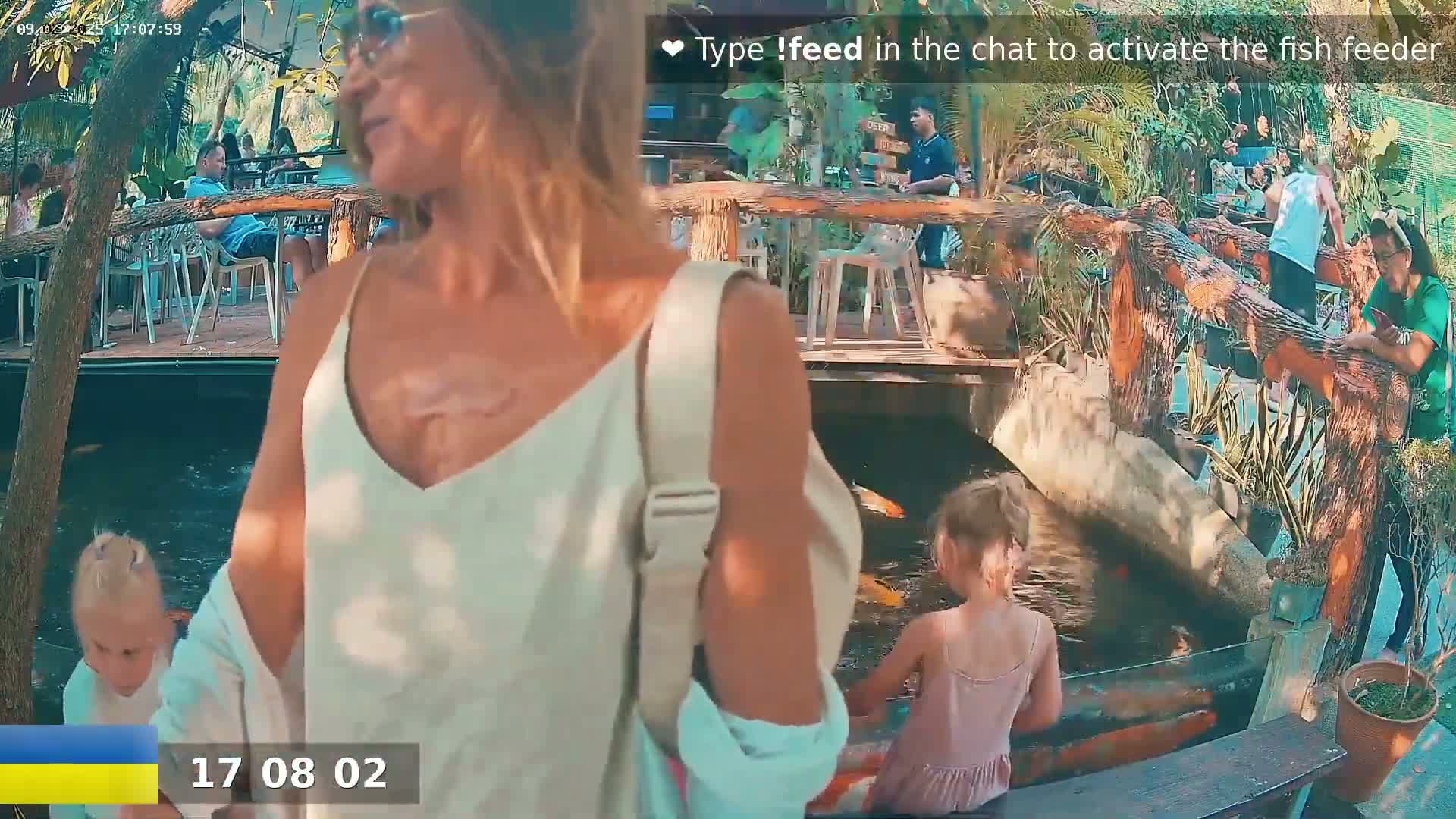 A woman and a young girl stand near a pond with colorful fish, looking at the fish, while other people sit at tables in an outdoor restaurant setting.
