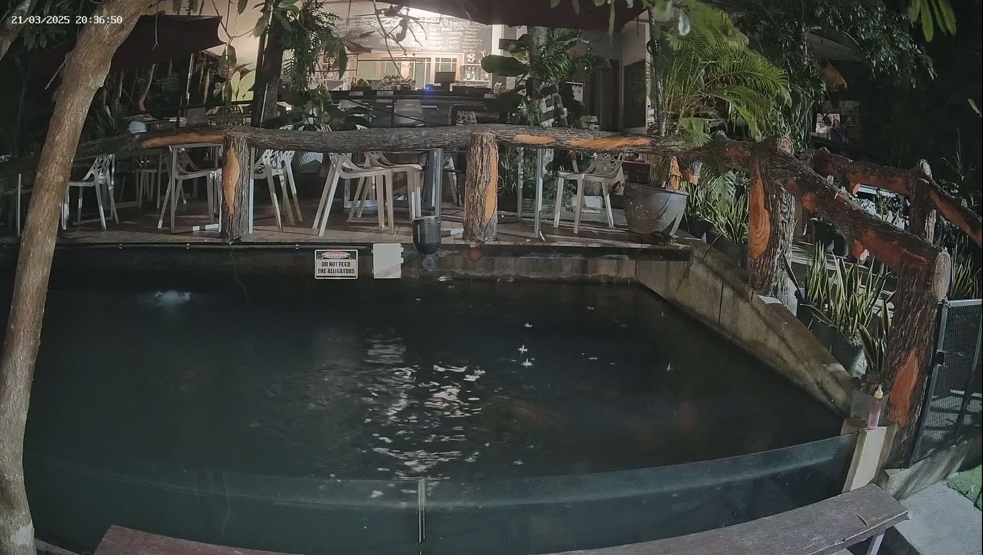At night, an outdoor patio area with empty tables and chairs overlooks a dark, rectangular pond.
