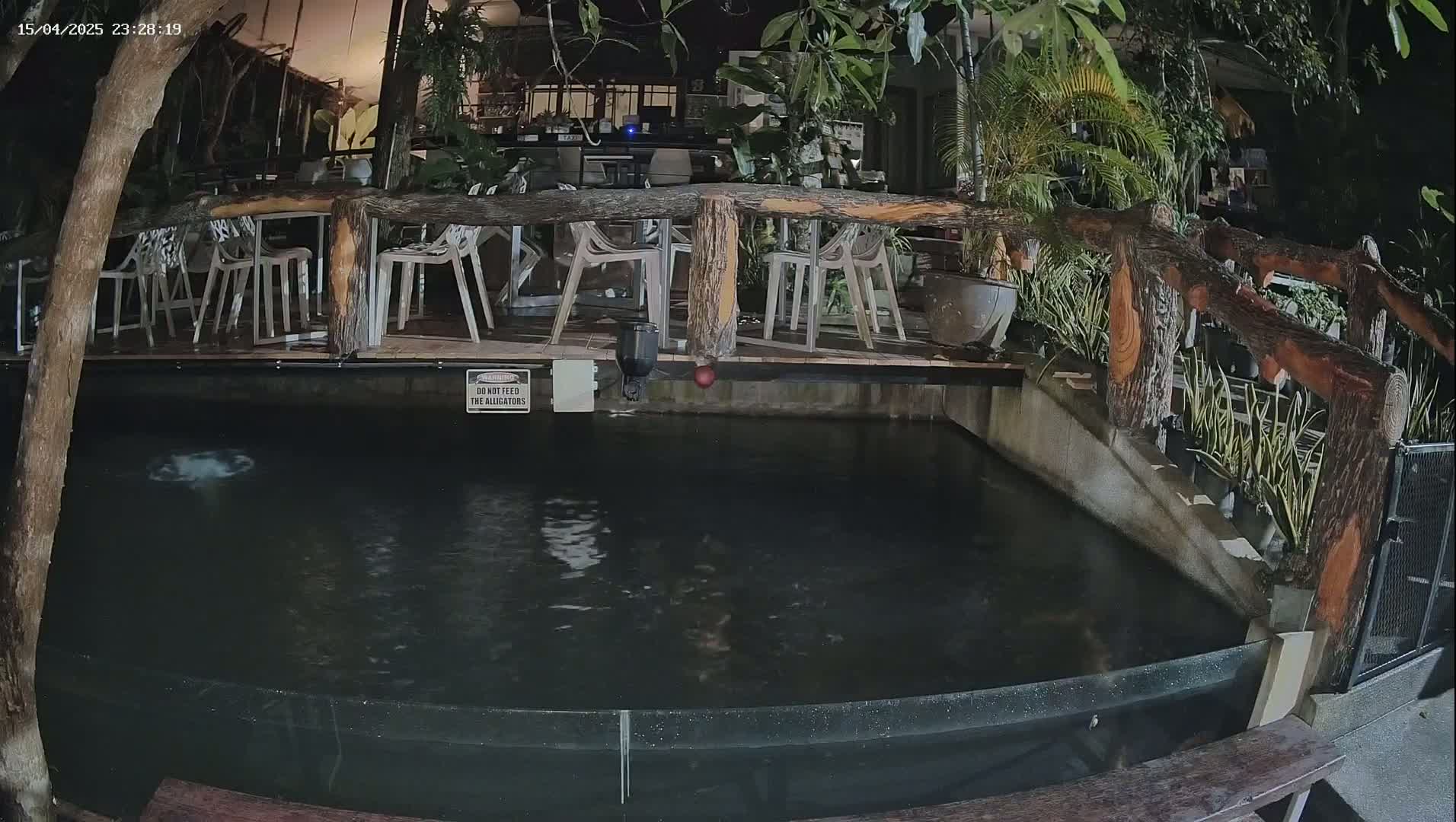 A dark, still pond is situated beneath a wooden deck with empty chairs and tables at night.