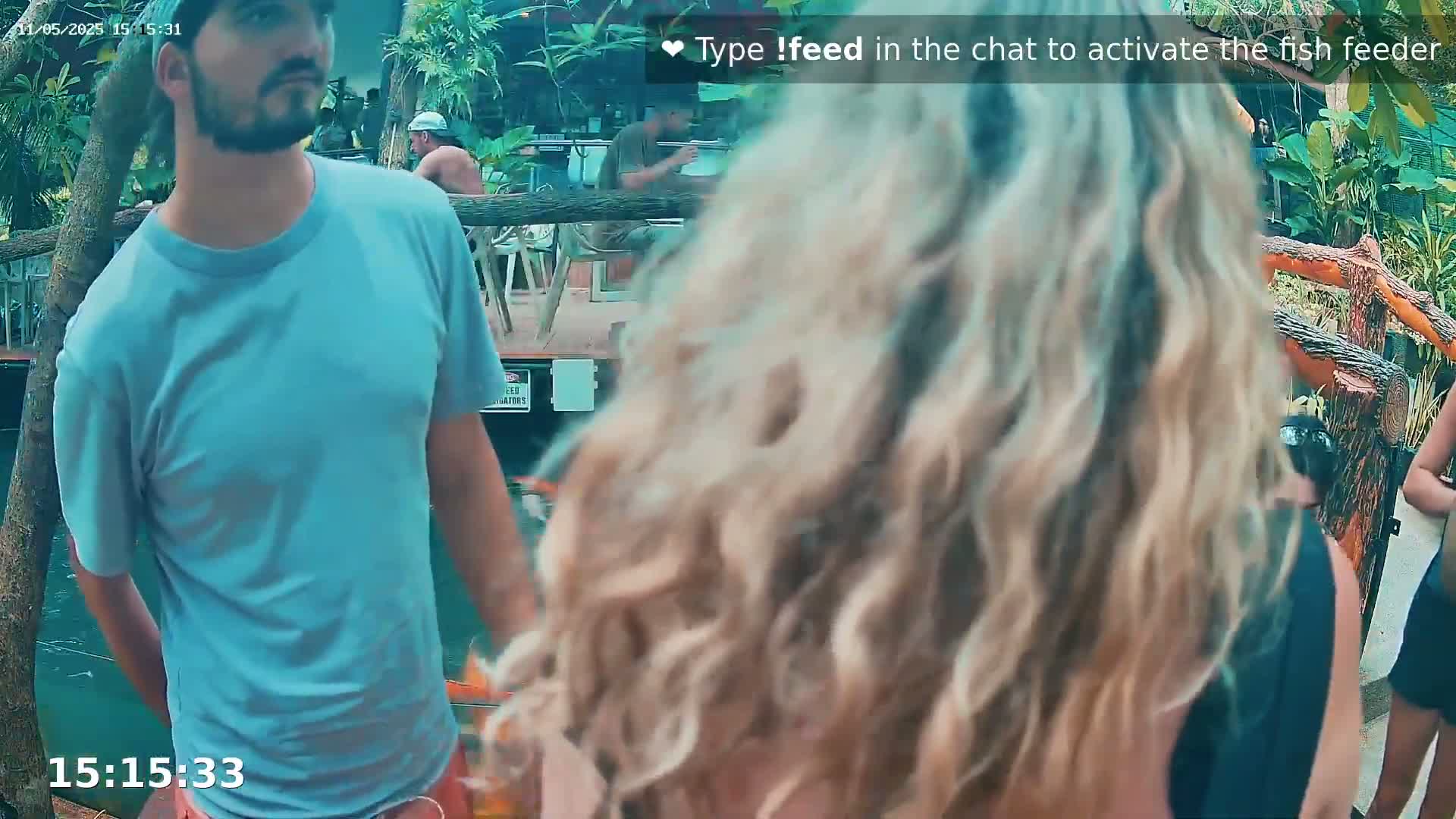 A man in a light blue shirt stands near a blonde woman with long, wavy hair, outdoors by a wooden railing near water.