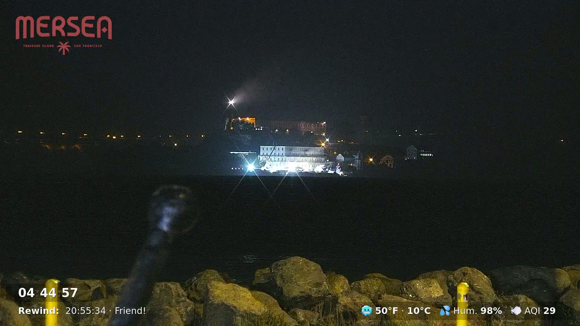 A nighttime view across dark water shows a brightly lit building complex on a distant hill, with a few lights visible on the shoreline.
