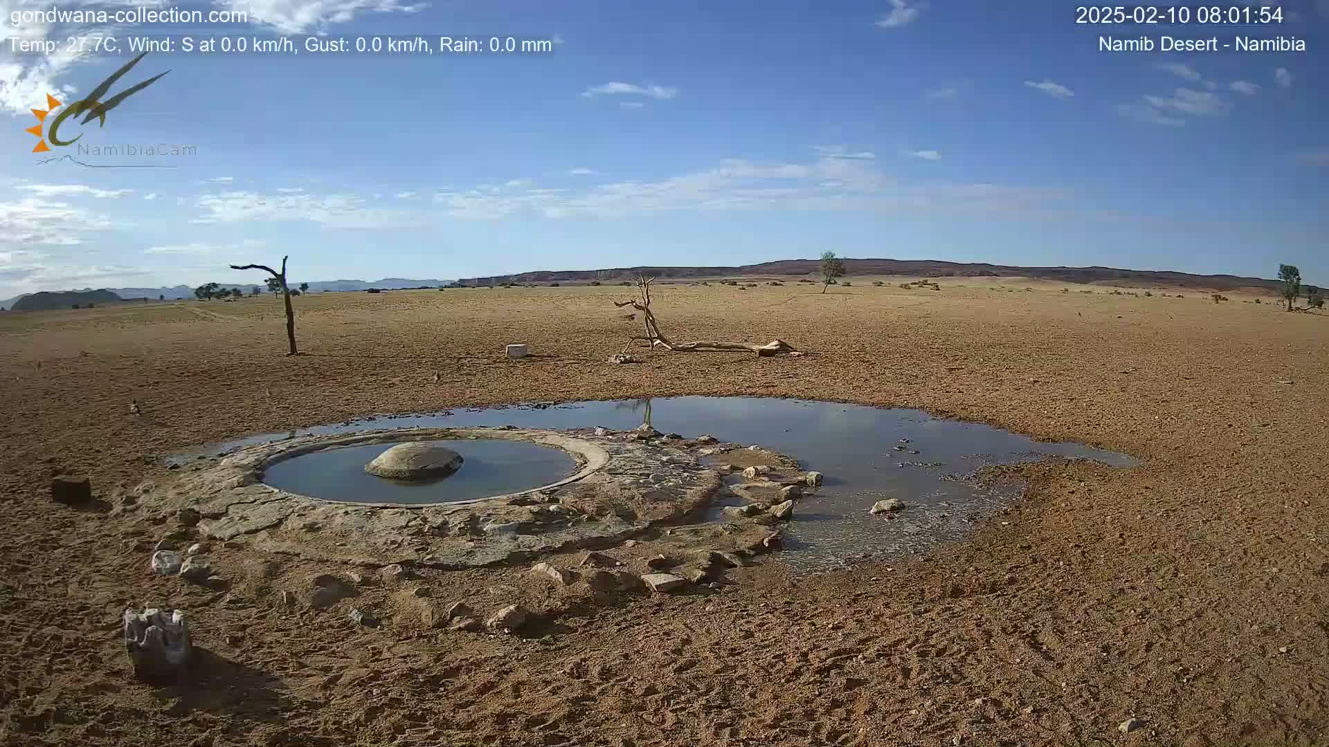 A small, shallow pool of water sits within a man-made stone structure in a dry, sandy desert under a mostly clear blue sky.