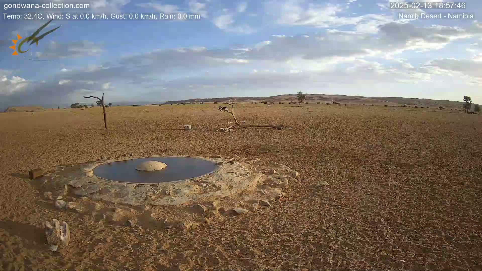 A small, circular pool of water sits in the center of a sandy desert landscape under a partly cloudy sky.