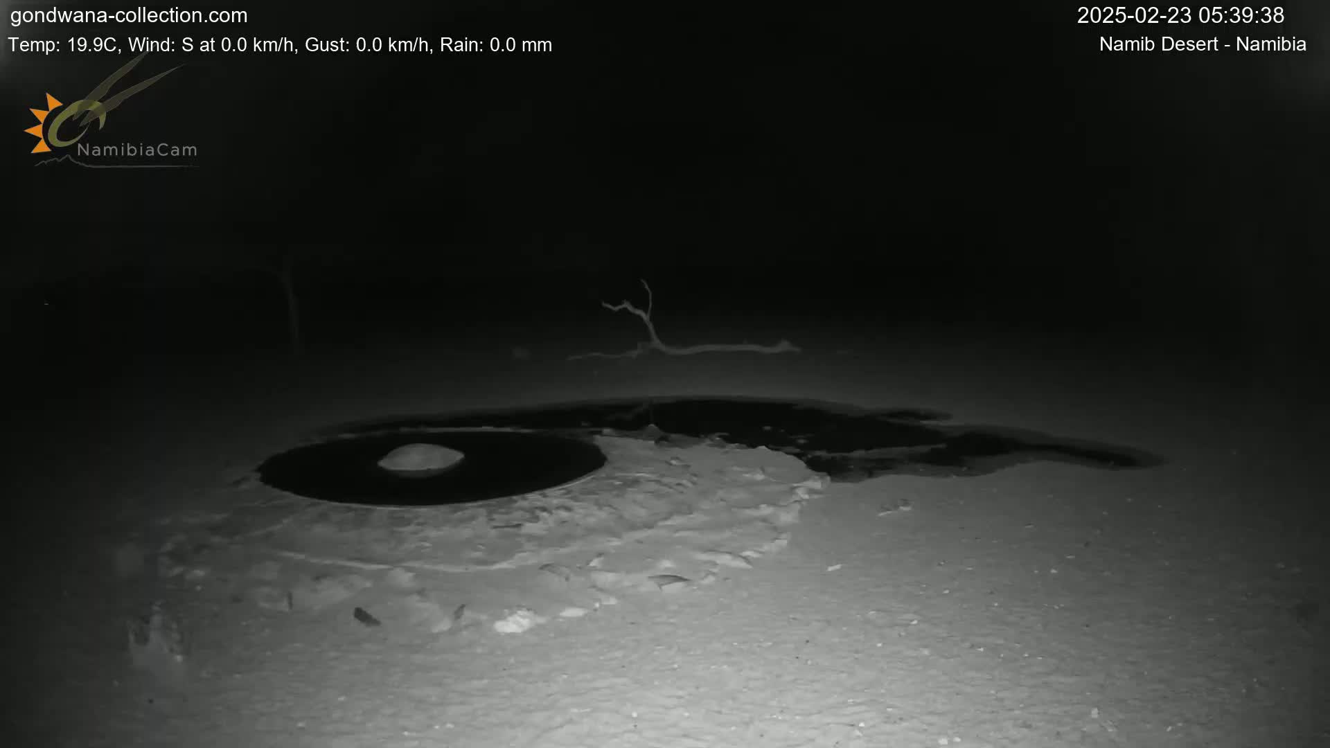 A dark, nighttime scene shows a circular, dark patch of water surrounded by lighter-colored sand or snow.