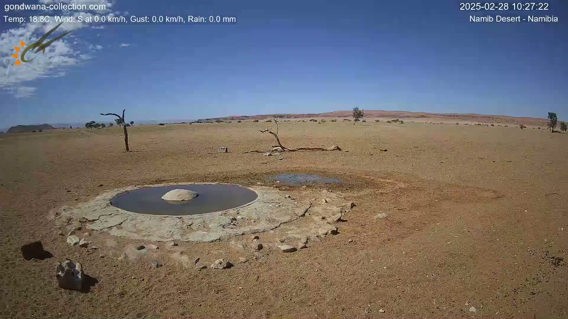 A small, shallow pool of water sits in the center of a dry, arid landscape under a clear blue sky.