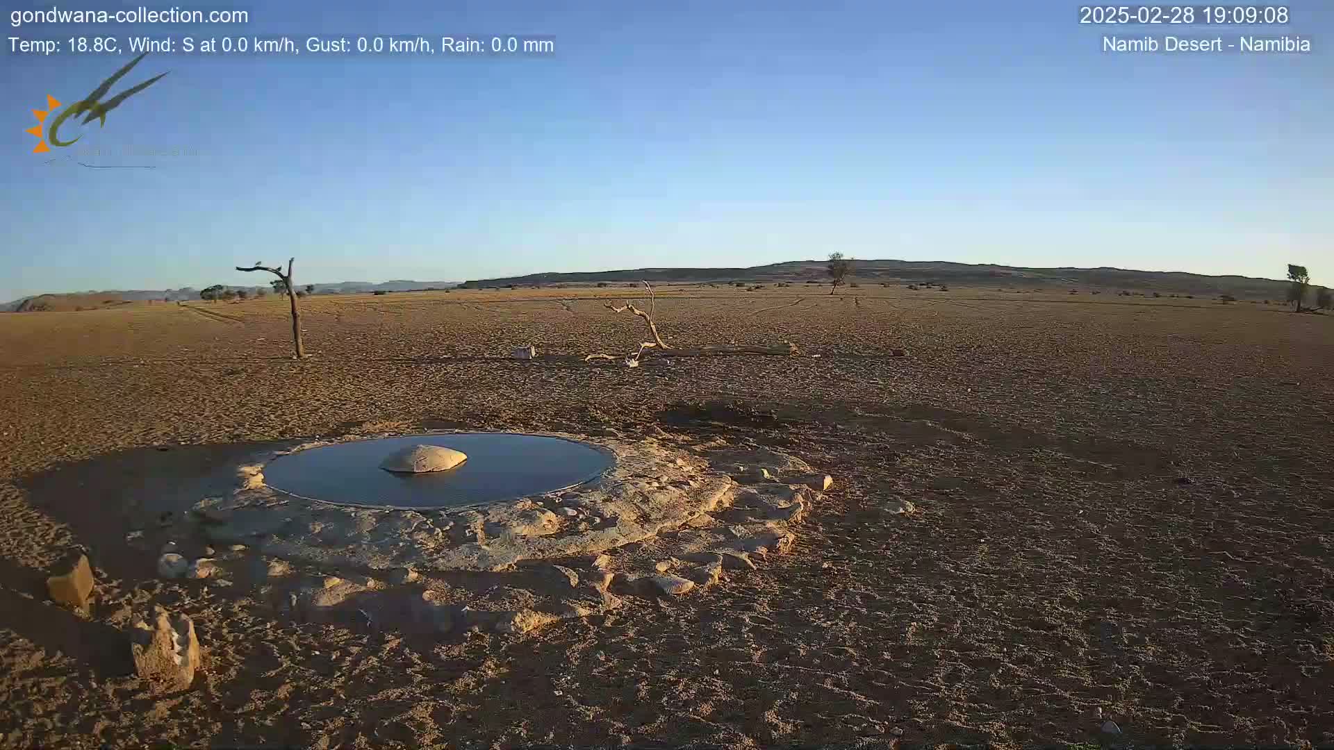 A small, circular pool of water sits in the center of a dry, sandy desert landscape under a clear blue sky.