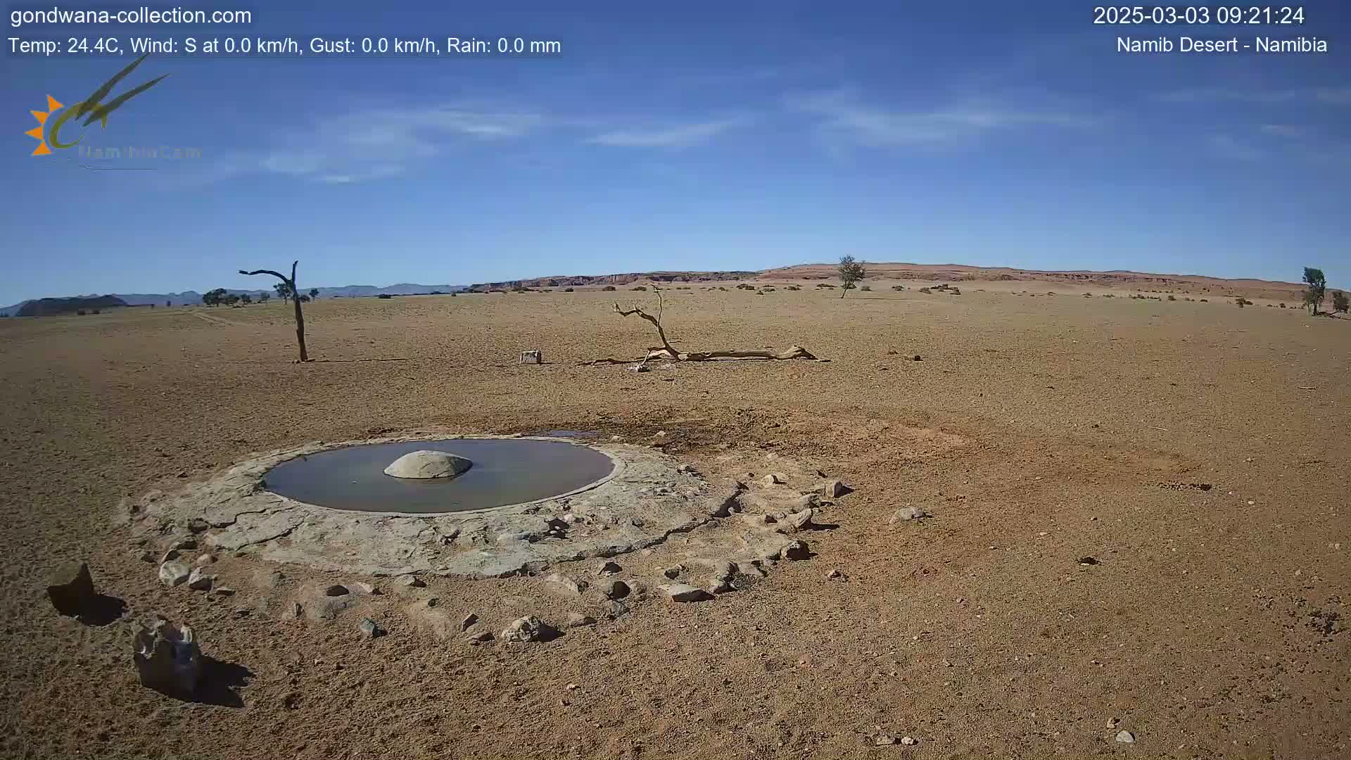 A small, shallow pool of water sits in a man-made basin in a dry, arid desert landscape under a clear blue sky.