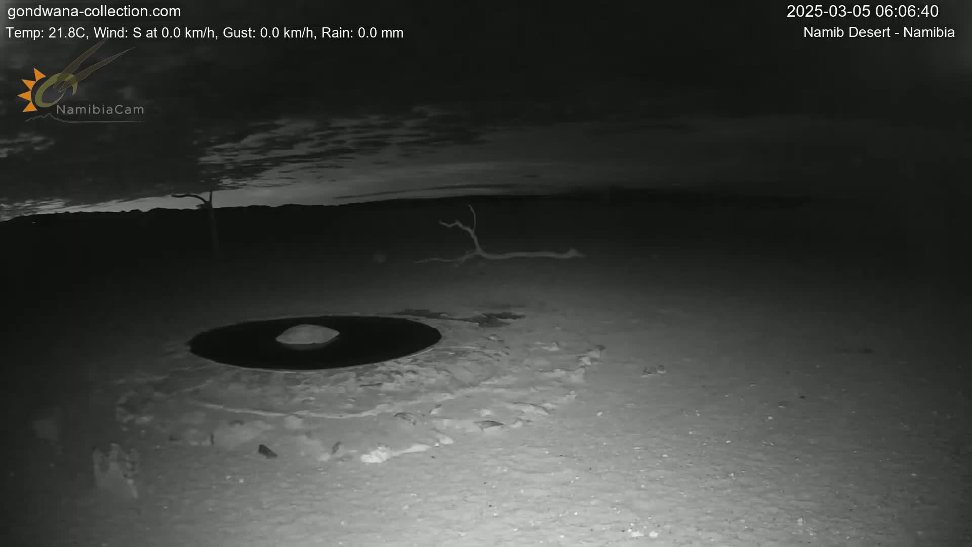 A dark, nighttime desert scene shows a circular depression in the sand with a small mound in the center, under a mostly clear sky.