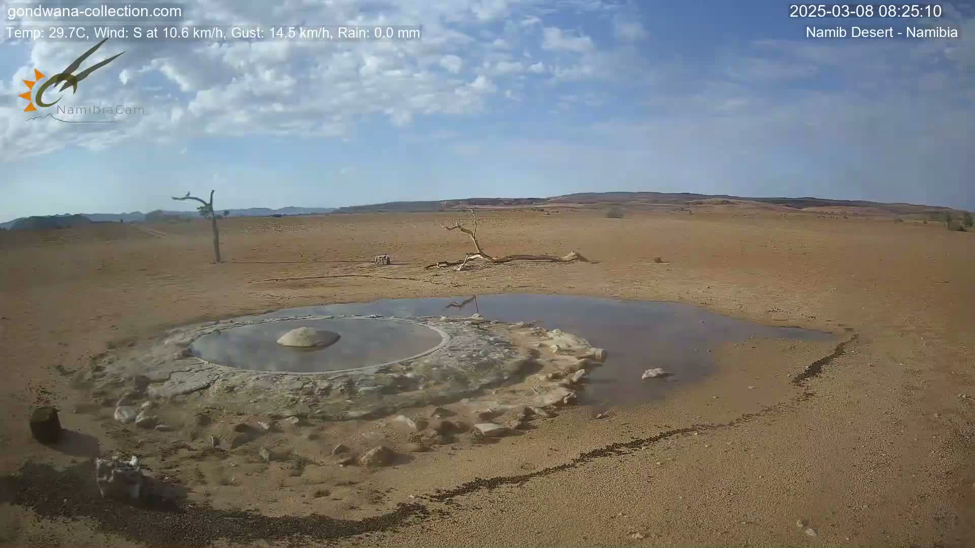 A mostly clear sky is above a desert landscape featuring a small, shallow pool of water surrounding a raised, circular structure of light-colored earth.
