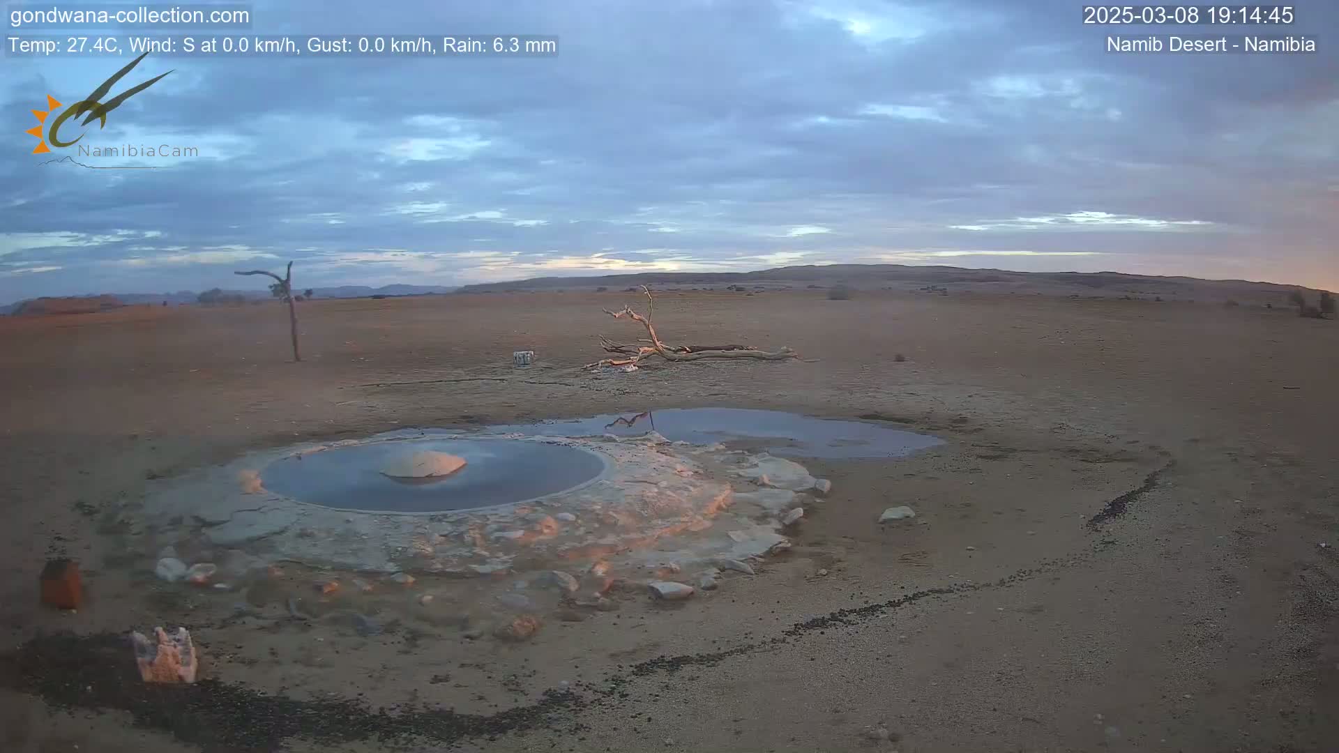 A desert landscape under a partly cloudy twilight sky shows a small, shallow pool of water next to a larger, circular pool surrounded by light-colored earth.