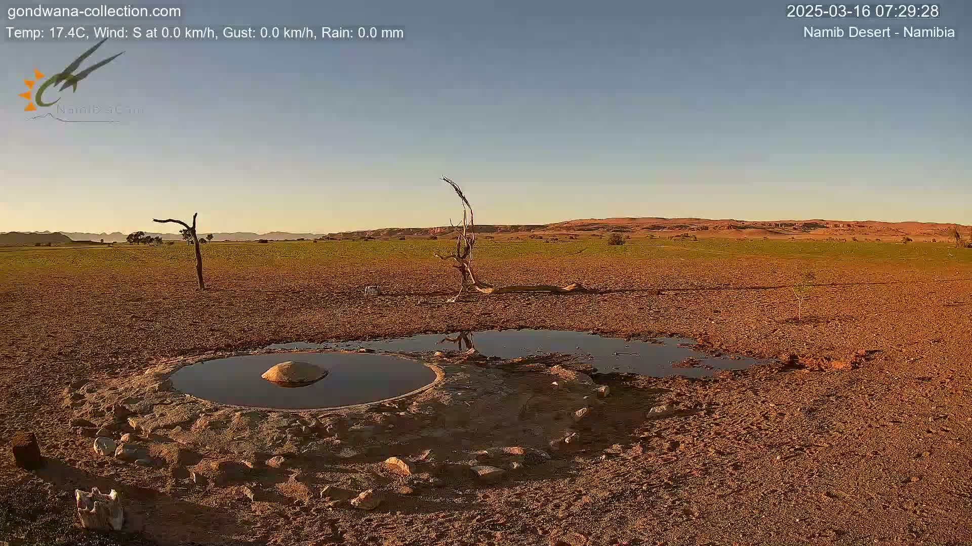 A small, circular pool of water sits in a dry, brown desert landscape under a clear, light-blue sky at sunrise.