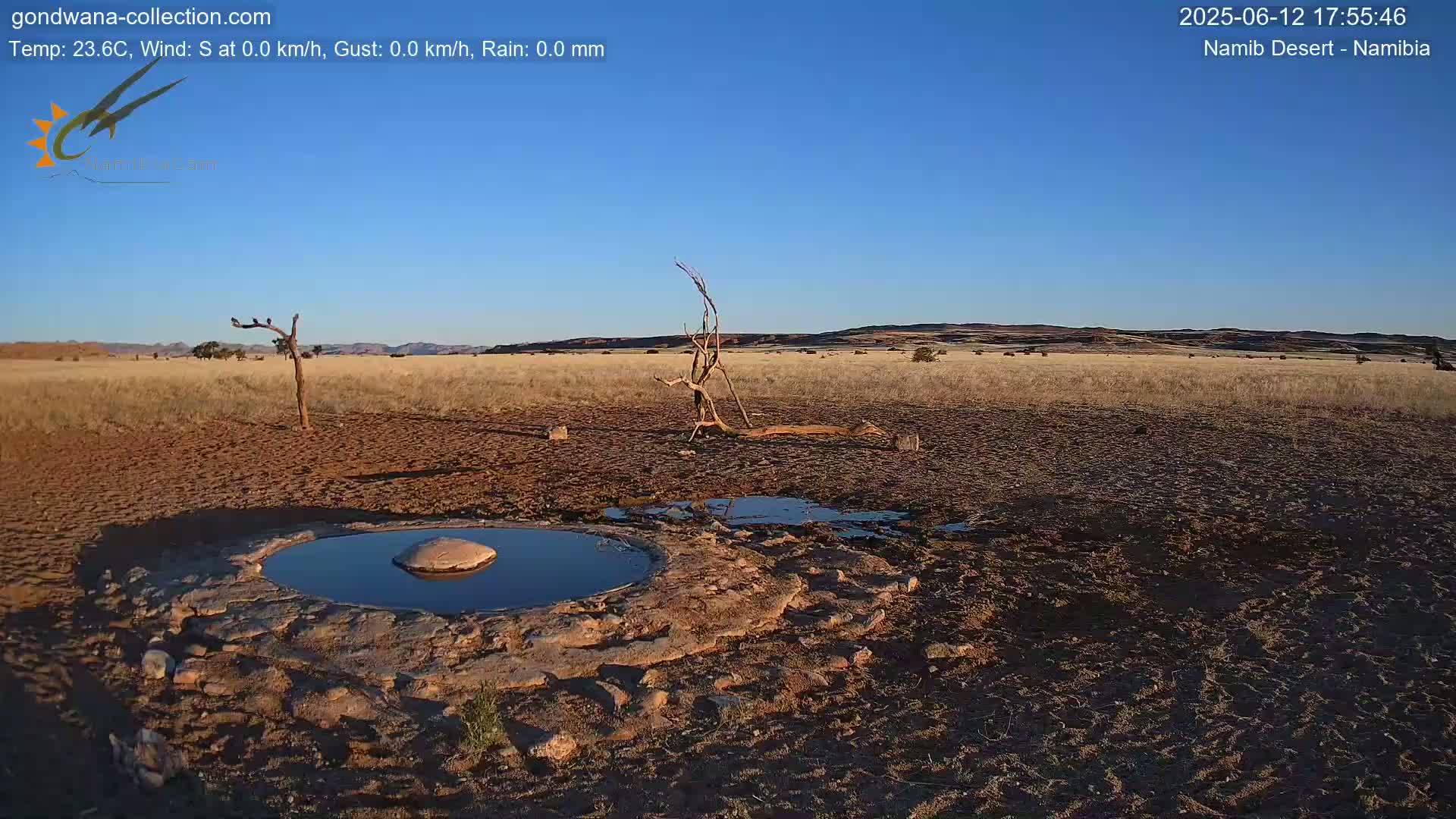 A small, still pool of water sits in a shallow, rocky basin in a dry, arid landscape under a clear blue sky.