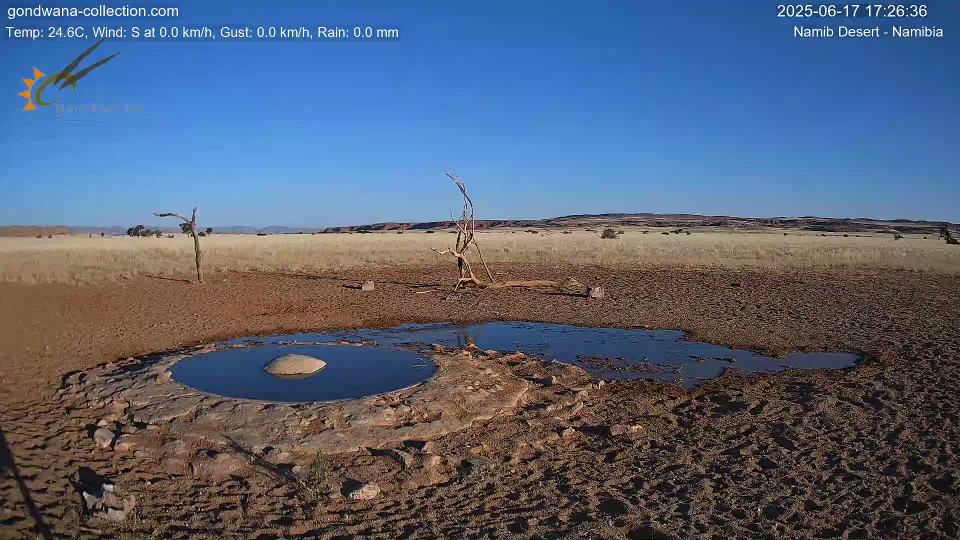 A small, dark pool of water sits in a dry, reddish-brown desert landscape under a clear blue sky.