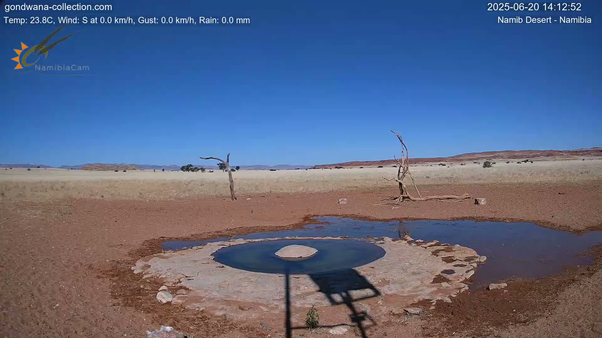 A small, shallow pool of water sits in a dry, reddish-brown landscape under a clear blue sky.
