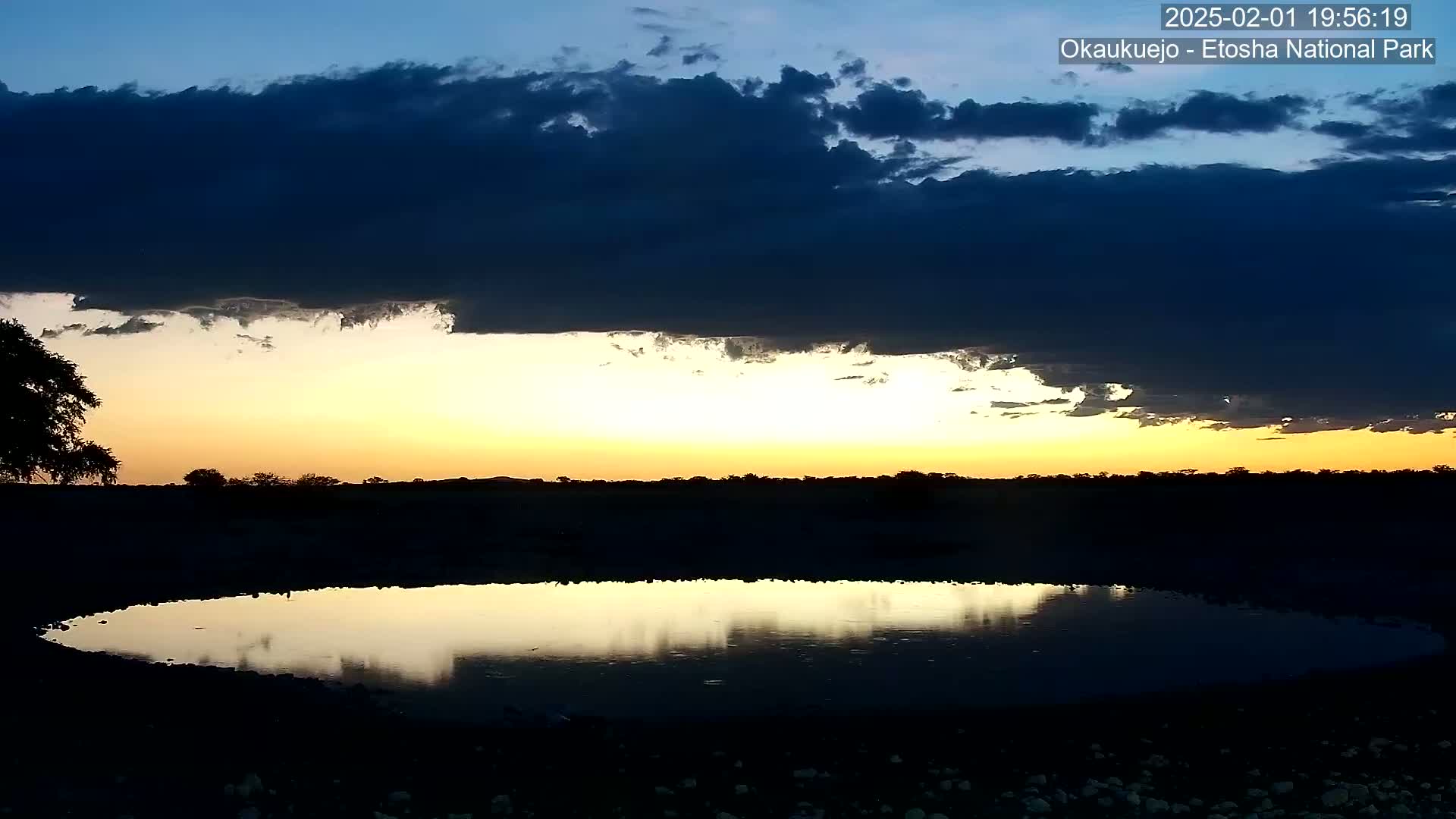 A still, dark-clouded sunset reflects in a calm, shallow pool of water in a flat, dark landscape.