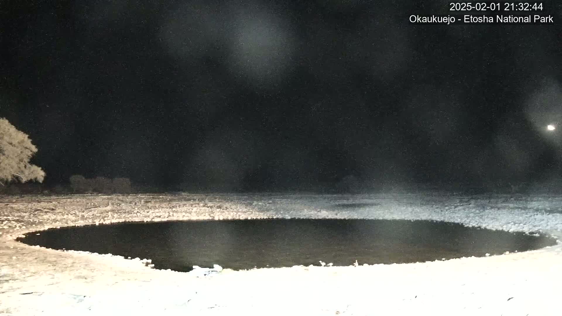 A dark, circular body of water is surrounded by a snow-covered landscape under a nighttime sky with light snowfall.