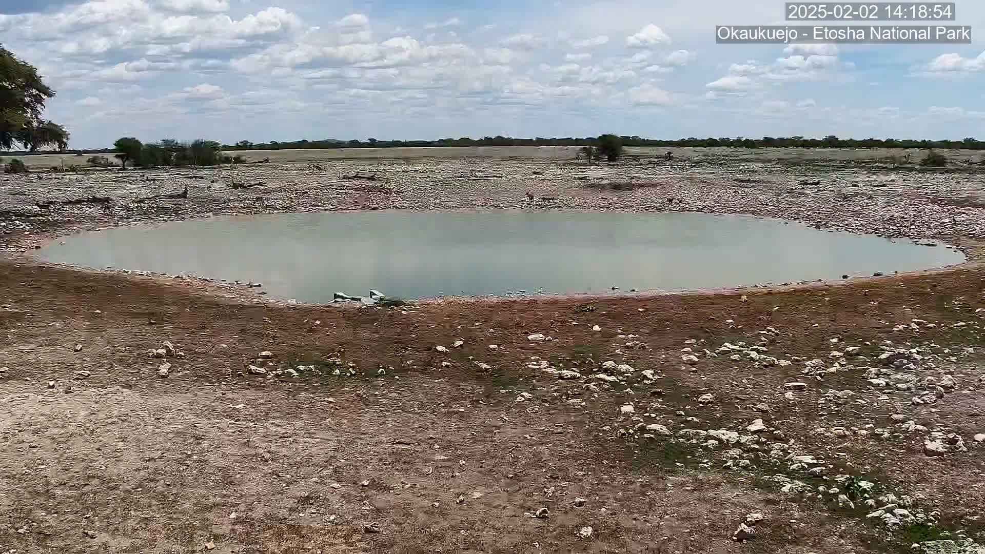 A mostly cloudy sky overlooks a small, calm body of water surrounded by rocky, dry land with sparse vegetation.