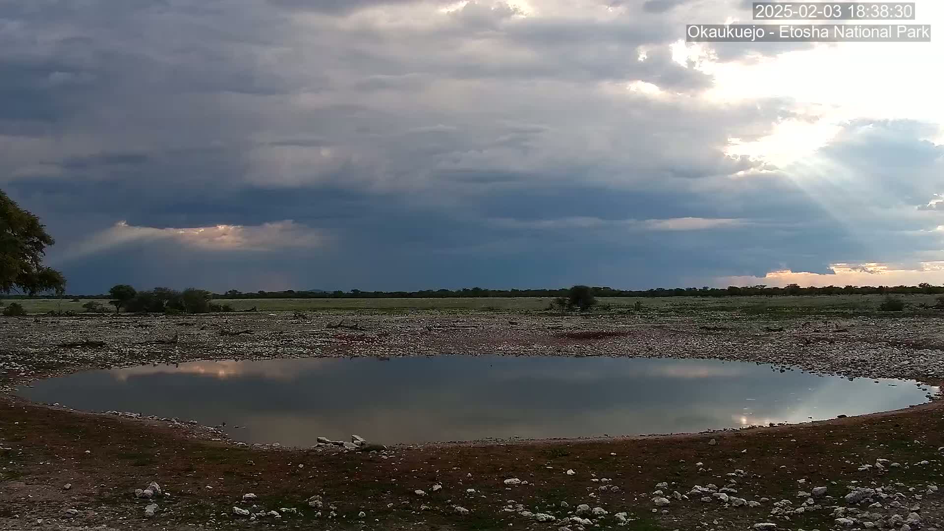 A small, still body of water reflects a partly cloudy sky with sunbeams breaking through the clouds in a rocky, sparsely vegetated landscape.