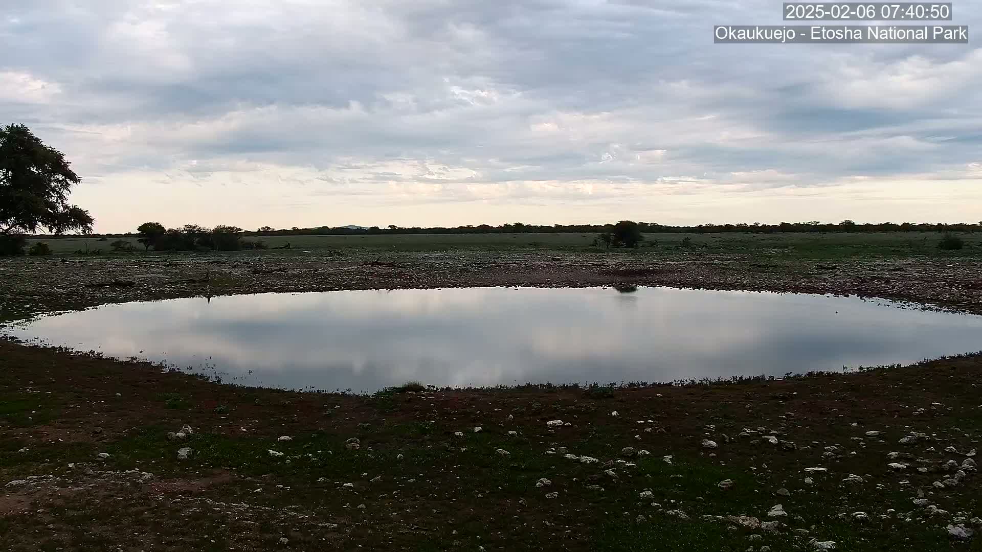A calm, still pool of water reflects a cloudy sky in a dry, rocky landscape.