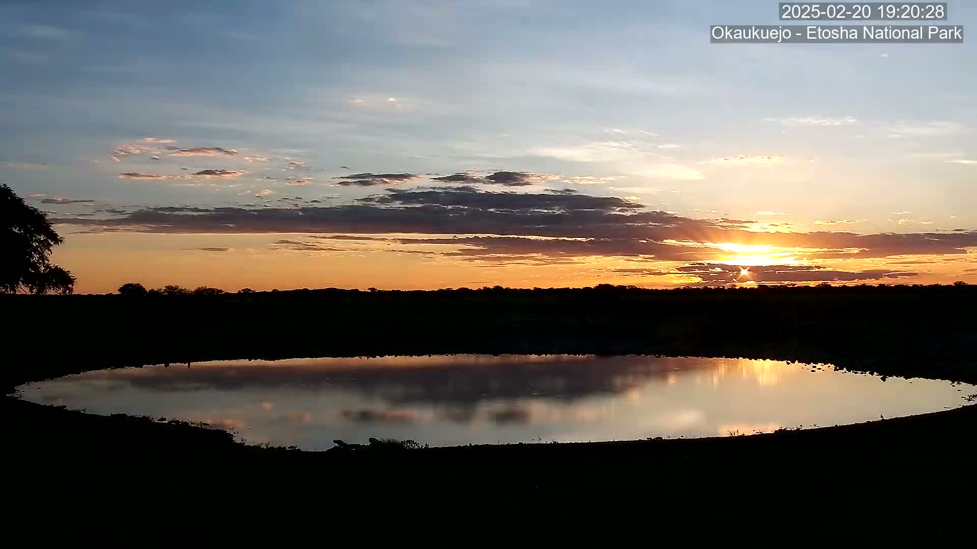 A calm body of water reflects a sunset with orange and yellow hues in the sky, partially obscured by clouds, over a silhouetted landscape.