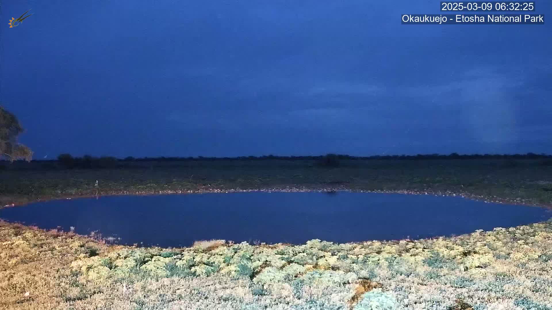 A dark blue, still body of water sits in a flat, dry landscape under a twilight sky.
